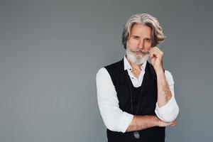 contra un fondo gris. un anciano moderno y elegante con pelo gris y barba está en el interior foto