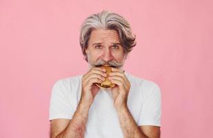 Eats delicious burger. Stylish modern senior man with gray hair and beard is indoors photo