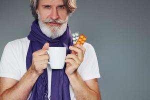 Getting sick. Against grey background. Stylish modern senior man with gray hair and beard is indoors photo