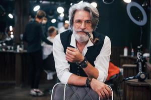 In retro clothes in barbershop. Stylish modern senior man with gray hair and beard is indoors photo