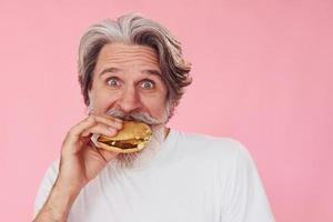 Eats delicious burger. Stylish modern senior man with gray hair and beard is indoors photo