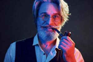 Smoking and having fun. Neon lighting. Stylish modern senior man with gray hair and beard is indoors photo