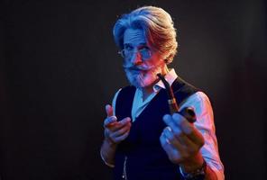 Smoking and having fun. Neon lighting. Stylish modern senior man with gray hair and beard is indoors photo