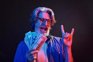 Holding cash. Neon lighting. Stylish modern senior man with gray hair and beard is indoors photo