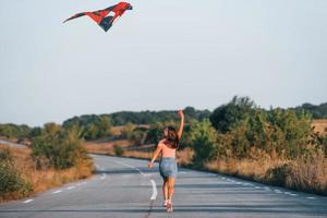 joven mujer alegre divirtiéndose con cometa al aire libre en la carretera foto