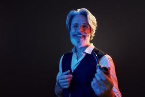 Smoking and having fun. Neon lighting. Stylish modern senior man with gray hair and beard is indoors photo