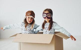 Sitting in the paper box. Two cute little girls indoors at home together. With retro pilot glasses and hat photo