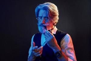 Smoking and having fun. Neon lighting. Stylish modern senior man with gray hair and beard is indoors photo