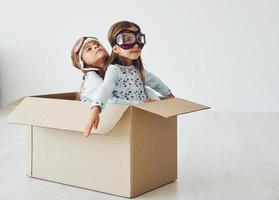 Sitting in the paper box. Two cute little girls indoors at home together. With retro pilot glasses and hat photo