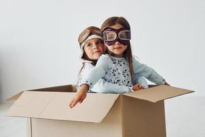 Sitting in the paper box. Two cute little girls indoors at home together. With retro pilot glasses and hat photo