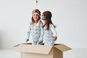 Sitting in the paper box. Two cute little girls indoors at home together. With retro pilot glasses and hat photo