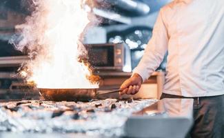 Frying pan is on fire. Chef in white uniform cooking food at kitchen. Busy day at work photo