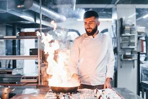 Frying pan is on fire. Chef in white uniform cooking food at kitchen. Busy day at work photo