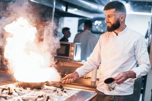 Frying pan is on fire. Chef in white uniform cooking food at kitchen. Busy day at work photo