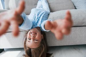 Lying upside down on the sofa. Cute little girl indoors at home alone. Child enjoying weekend photo