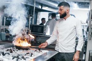 Frying pan is on fire. Chef in white uniform cooking food at kitchen. Busy day at work photo