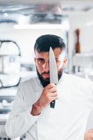 Chef in white uniform standing at kitchen. Holding knife in hands photo