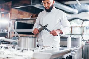 Chef in white uniform standing at kitchen. Holding knives in hands photo