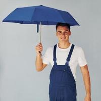 sostiene el paraguas a mano. trabajador de sexo masculino en uniforme azul de pie dentro del estudio contra el fondo blanco foto