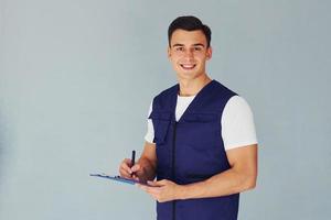 Writes in notepad. Male worker in blue uniform standing inside of studio against white background photo