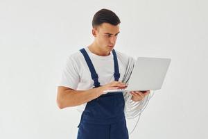 utiliza portátil. trabajador de sexo masculino en uniforme azul de pie dentro del estudio contra el fondo blanco foto