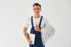 Holds cables, notepad and screwdriver. Male worker in blue uniform standing inside of studio against white background photo