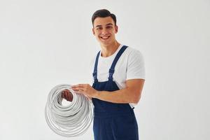 sostiene cable. trabajador de sexo masculino en uniforme azul de pie dentro del estudio contra el fondo blanco foto