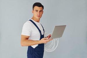 Uses laptop. Male worker in blue uniform standing inside of studio against white background photo