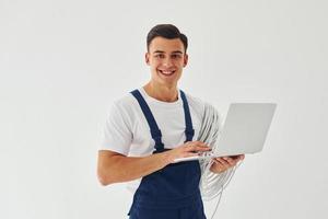 utiliza portátil. trabajador de sexo masculino en uniforme azul de pie dentro del estudio contra el fondo blanco foto