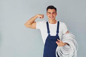 sostiene cable. trabajador de sexo masculino en uniforme azul de pie dentro del estudio contra el fondo blanco foto