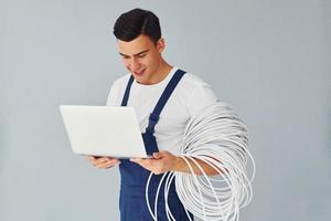 Uses laptop. Male worker in blue uniform standing inside of studio against white background photo