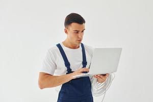 Uses laptop. Male worker in blue uniform standing inside of studio against white background photo