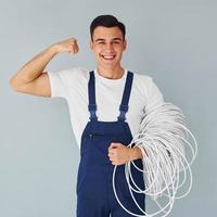 sostiene cable. trabajador de sexo masculino en uniforme azul de pie dentro del estudio contra el fondo blanco foto