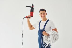 con taladro y cables en las manos. trabajador de sexo masculino en uniforme azul de pie dentro del estudio contra el fondo blanco foto