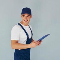 In cap and with notepad. Male worker in blue uniform standing inside of studio against white background photo