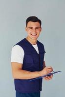 Writes in notepad. Male worker in blue uniform standing inside of studio against white background photo