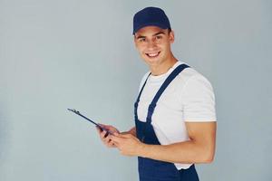 In cap and with notepad. Male worker in blue uniform standing inside of studio against white background photo