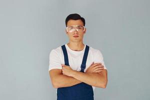 en gafas protectoras. brazos cruzados. trabajador de sexo masculino en uniforme azul de pie dentro del estudio contra el fondo blanco foto