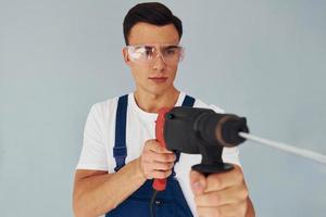 In protective eyewear and with drill in hands. Male worker in blue uniform standing inside of studio against white background photo