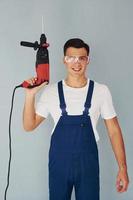 In protective eyewear and with drill in hands. Male worker in blue uniform standing inside of studio against white background photo