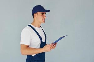 In cap and with notepad. Male worker in blue uniform standing inside of studio against white background photo