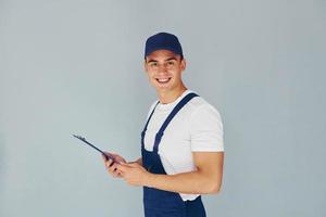 en gorra y con bloc de notas. trabajador de sexo masculino en uniforme azul de pie dentro del estudio contra el fondo blanco foto