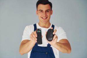 TV remote controller and computer mouse. Male worker in blue uniform standing inside of studio against white background photo