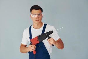 In protective eyewear and with drill in hands. Male worker in blue uniform standing inside of studio against white background photo