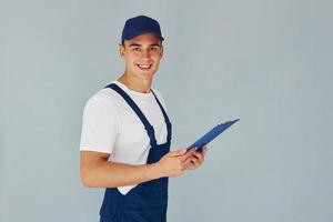 In cap and with notepad. Male worker in blue uniform standing inside of studio against white background photo