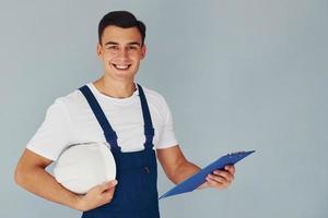 con bloc de notas y casco. trabajador de sexo masculino en uniforme azul de pie dentro del estudio contra el fondo blanco foto