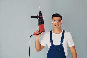 In protective eyewear and with drill in hands. Male worker in blue uniform standing inside of studio against white background photo