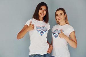 Two young women in shirts with heart shaped print on it standing indoors and shows thumbs up photo