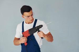 In protective eyewear and with drill in hands. Male worker in blue uniform standing inside of studio against white background photo