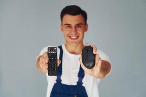 TV remote controller and computer mouse. Male worker in blue uniform standing inside of studio against white background photo